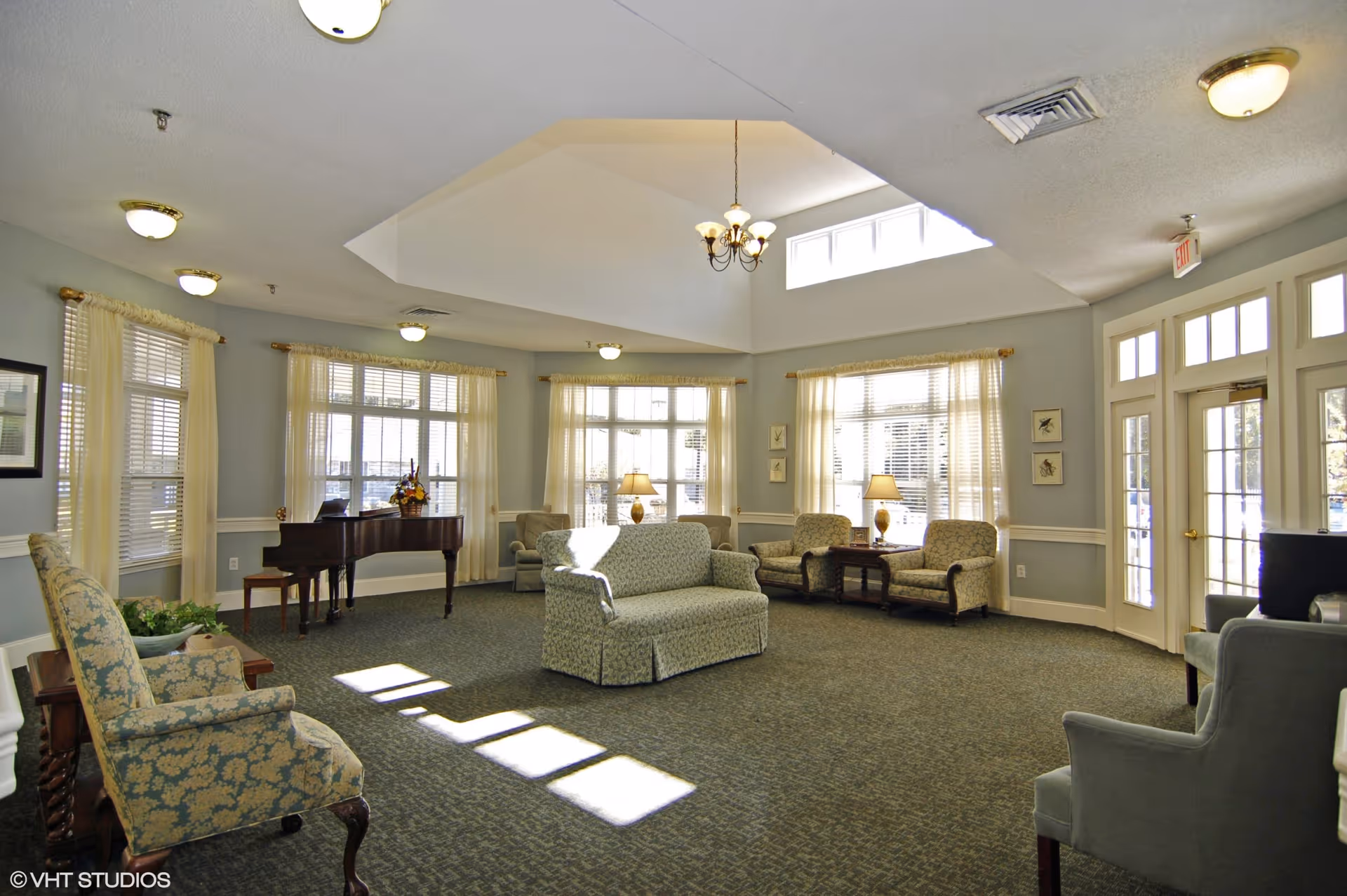 Bright, spacious senior living common room with seating, a grand piano, and large windows.