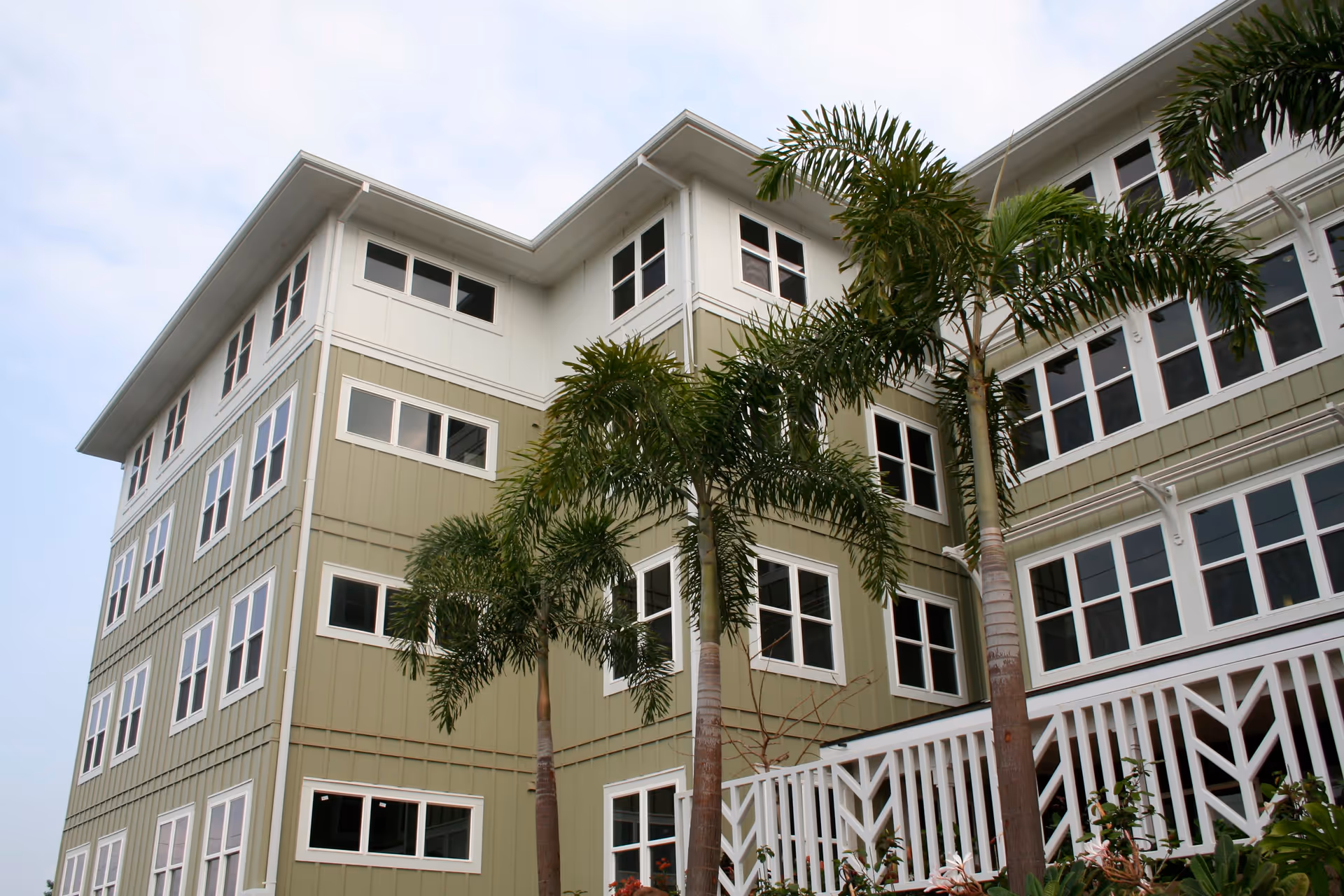 Exterior view of a multi-story senior living facility building with green and white siding, multiple windows, palm trees, and a white railing in front.
