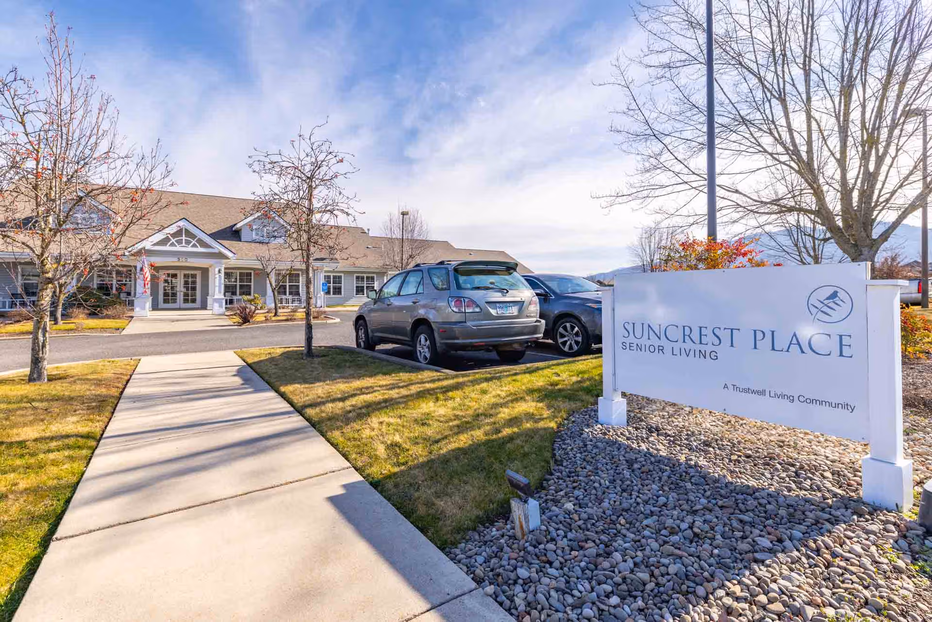 Walkway leads to Suncrest Place Senior Living exterior with entrance sign and parked cars.