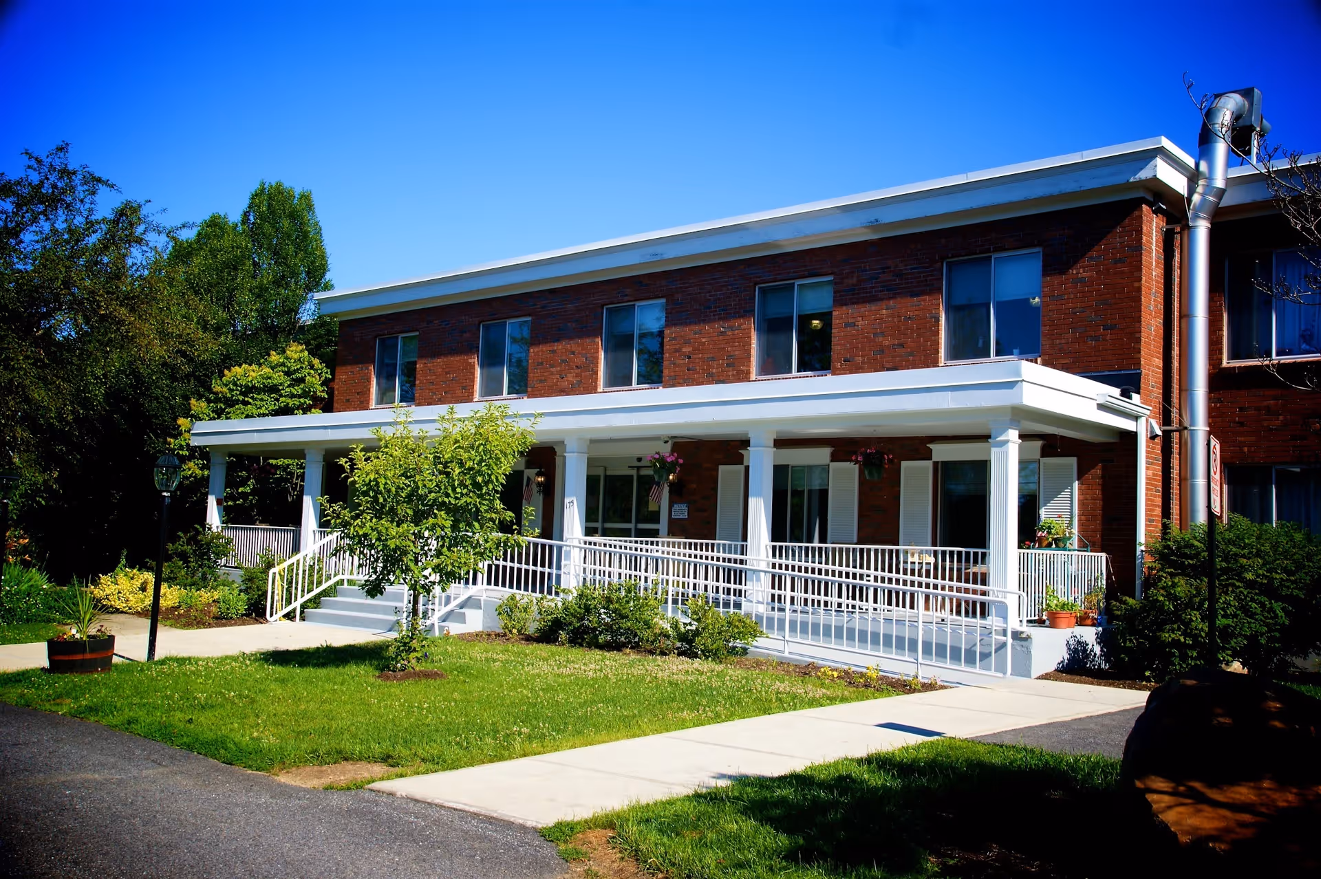 Two-story brick senior living building with a white covered porch, wheelchair ramp, and landscaped front lawn under a clear blue sky.