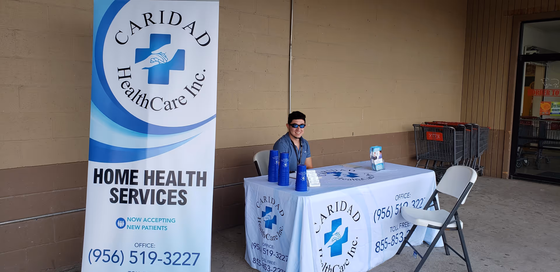 Outdoor promotional table for Caridad HealthCare Inc. with banner and staff member present.