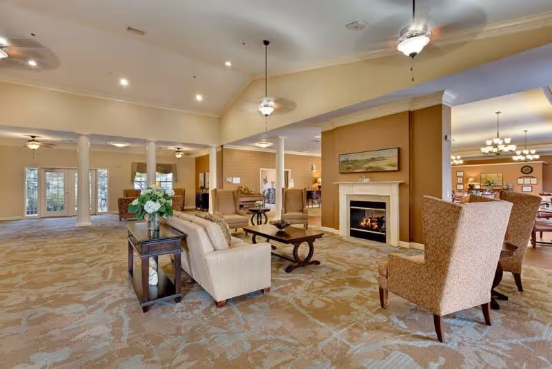 A spacious and well-lit living room area in a senior living facility featuring beige and brown tones. The room includes comfortable armchairs and sofas arranged around a coffee table and a fireplace with a painting above it. Ceiling fans and pendant lights hang from the high ceiling, and large windows and glass doors allow natural light to enter. In the background, there is a dining area with tables and chairs.