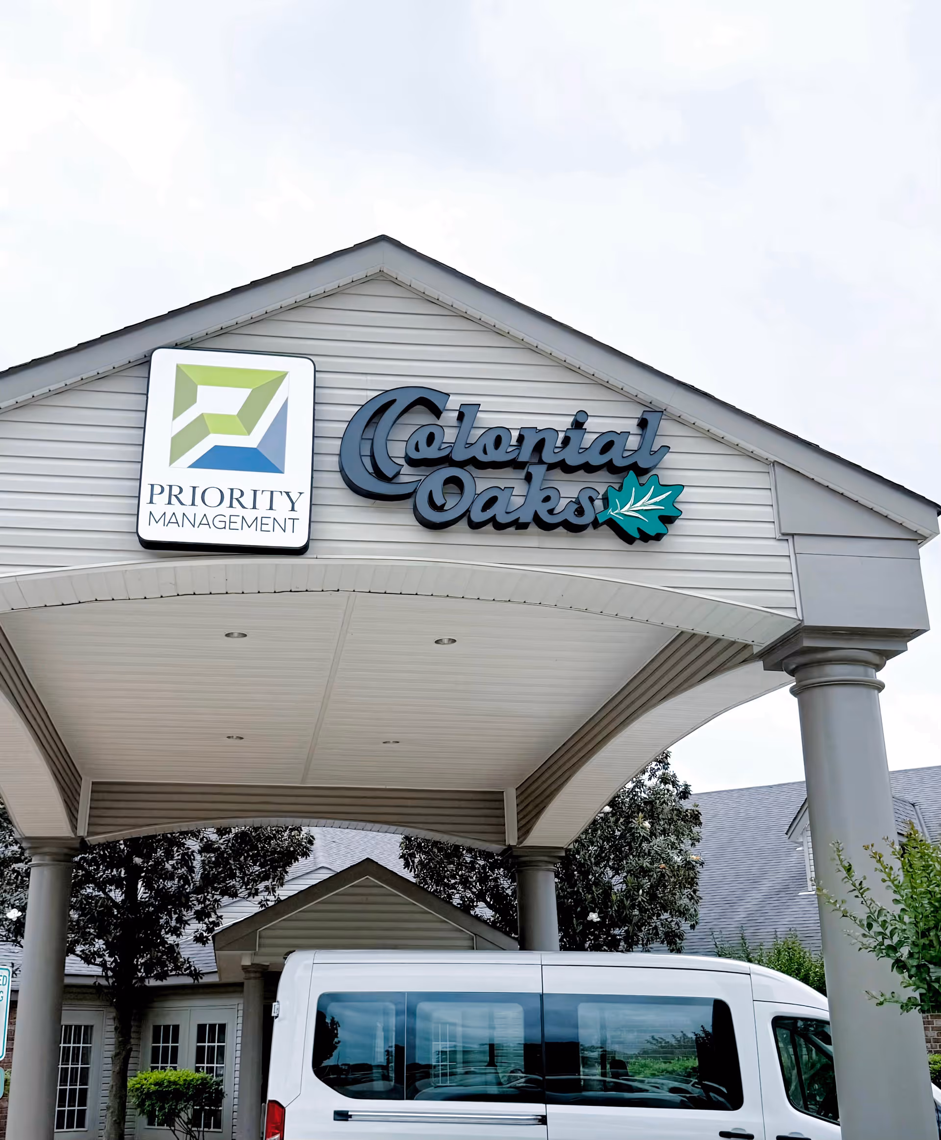 Entrance canopy of The Colonial Oaks Skilled Nursing & Rehabilitation facility with a white van parked underneath. The canopy has two signs: one for Priority Management and another with the Colonial Oaks logo featuring a green leaf. Trees and part of the building are visible in the background.
