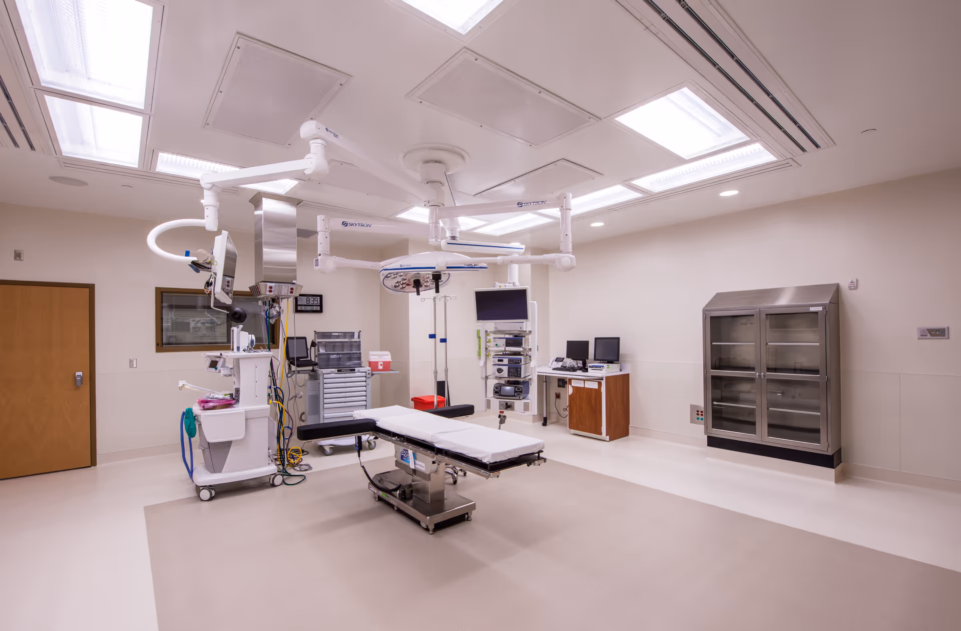 Sterile operating room with adjustable table, overhead lights, monitors, and cabinets.