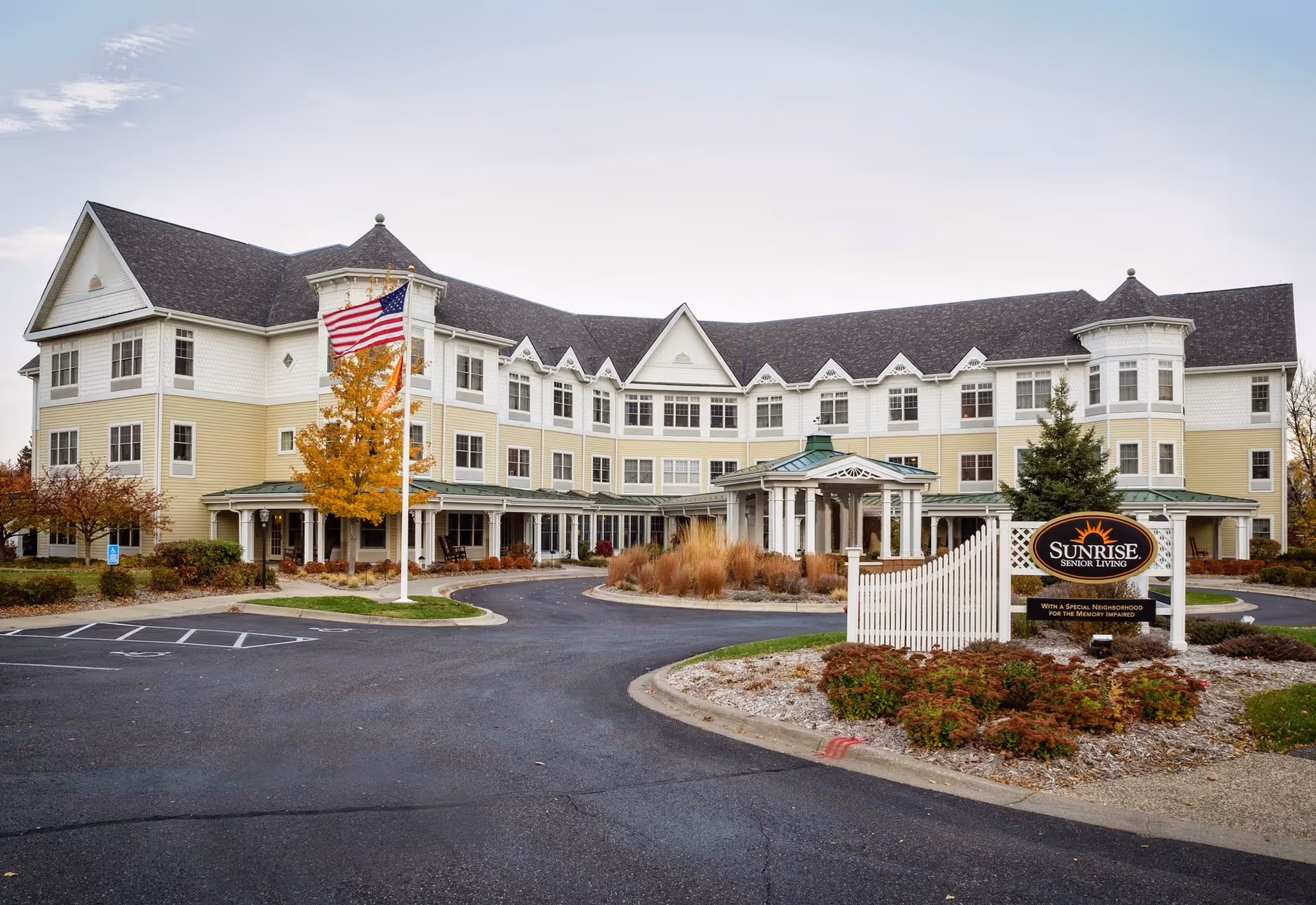 Exterior view of a large, three-story senior living facility with beige and white siding, multiple windows, and a covered entrance. There is an American flag on a flagpole and landscaped bushes and trees with autumn foliage around the building. A sign in front reads 'Sunrise Senior Living'.