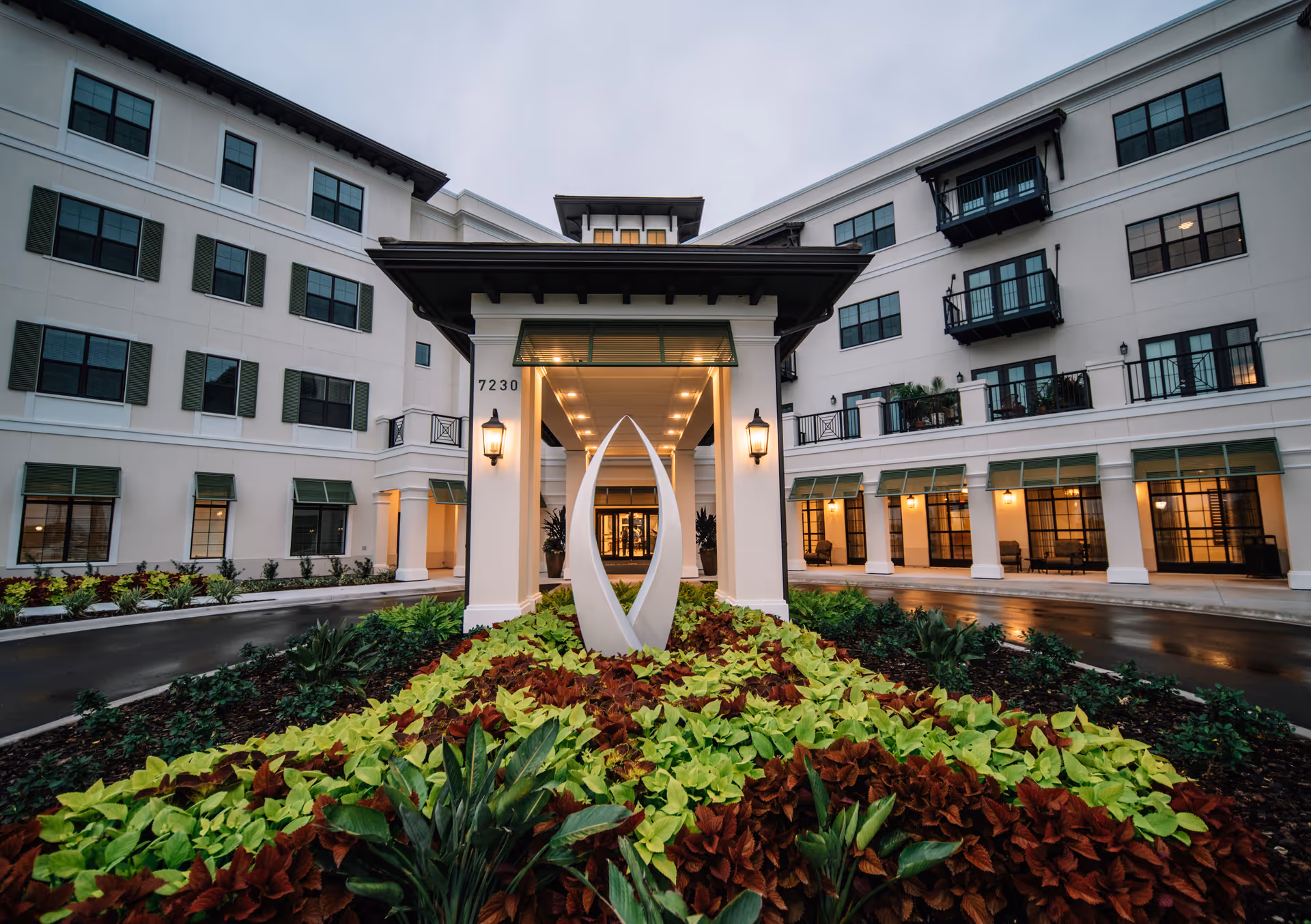Front entrance of a multi-story senior living building with a covered porte-cochere, modern sculpture, and landscaped flowerbeds.