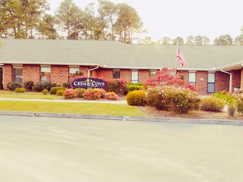 Single-story brick assisted living building with a 'Cedar Cove' sign, landscaped shrubs and an American flag in front.