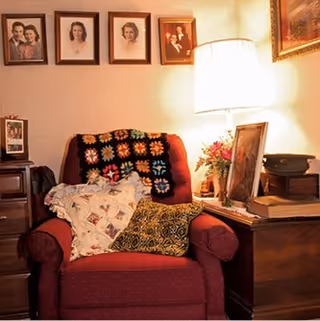 A cozy living room corner with a red armchair adorned with patterned pillows and a crocheted blanket. Next to the chair is a wooden side table holding a lamp, framed photos, flowers, and a vintage record player. Several framed family portraits hang on the wall above the chair.