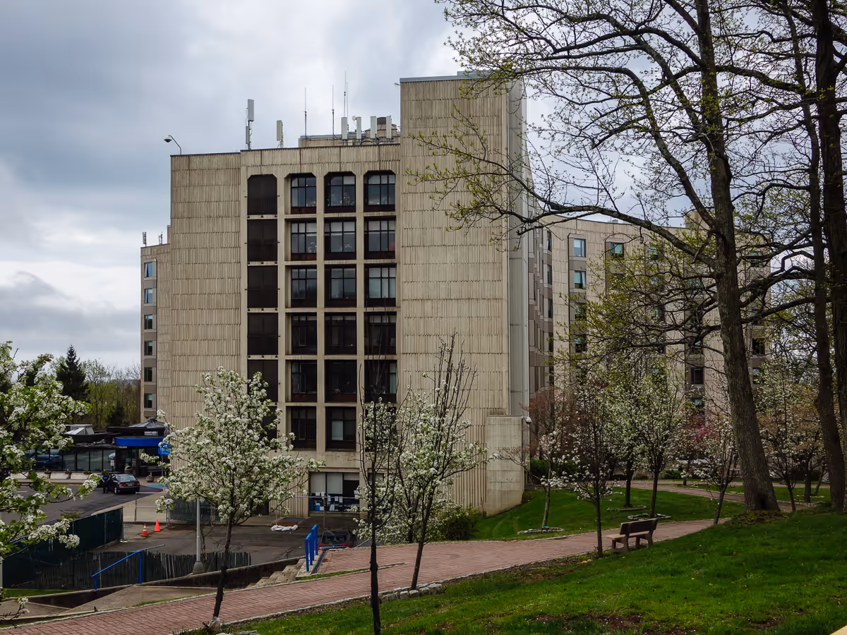 Front view of a tall beige building with trees and a bench in foreground.