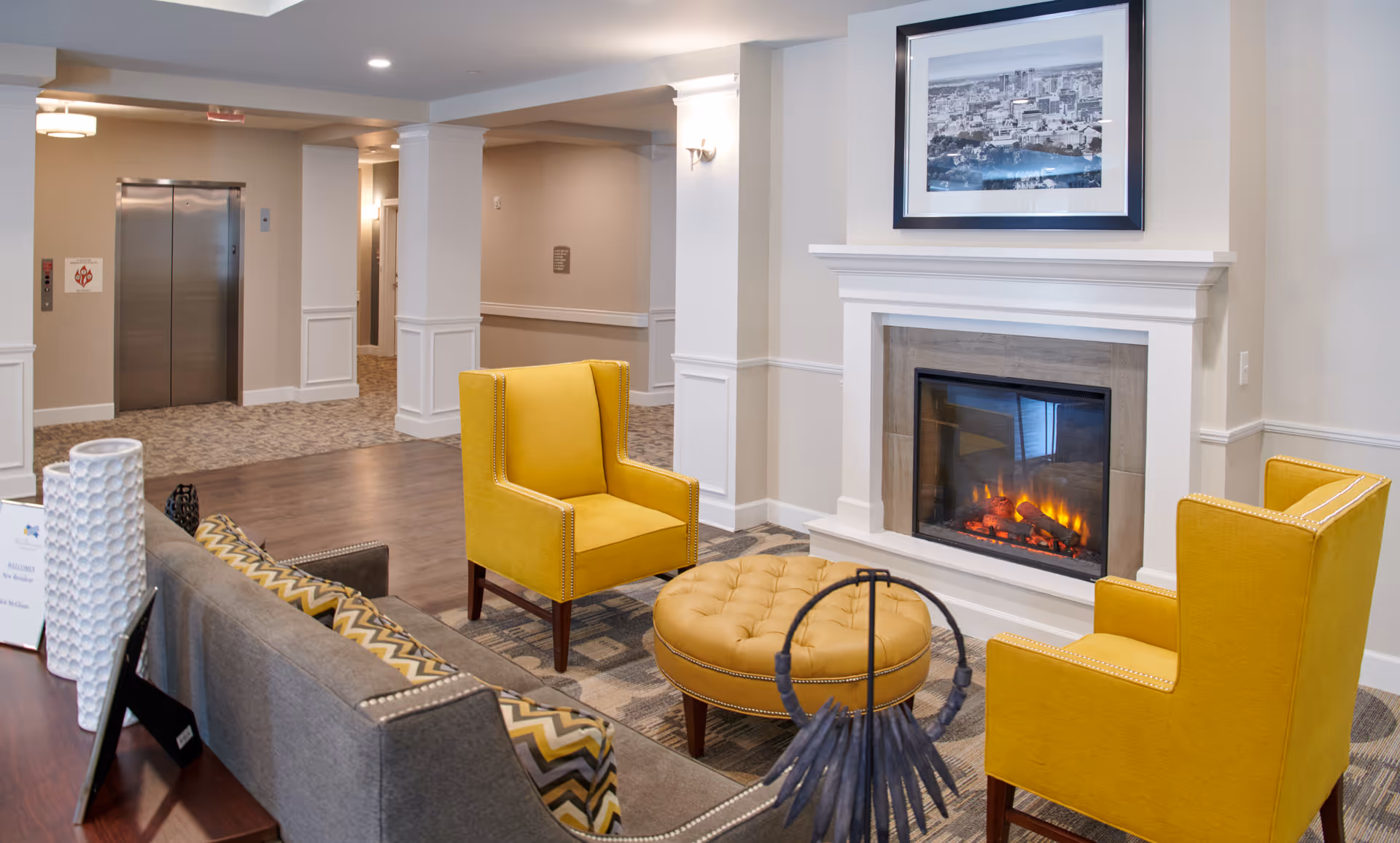 A cozy seating area in a senior living facility with two yellow armchairs, a round yellow ottoman, and a gray sofa with patterned pillows arranged around a lit fireplace. The room has neutral-colored walls, a framed black and white cityscape photo above the fireplace, and an elevator in the background.