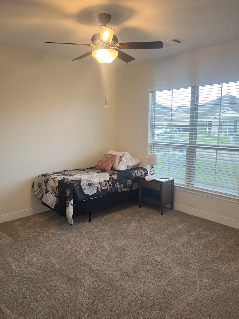 A furnished bedroom in a senior living facility featuring a bed, nightstand, lamp, and window.