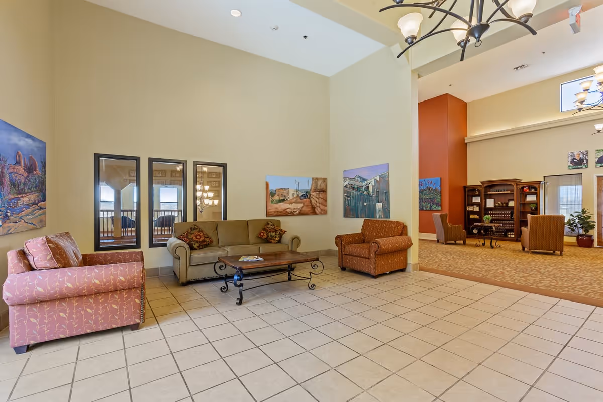 A spacious living area in Evergreen Village Prescott featuring a tiled floor, a beige sofa with patterned cushions, a pink armchair, and an orange armchair. The walls are decorated with colorful paintings and three vertical mirrors. In the background, there is a carpeted area with additional seating, a wooden cabinet, and large windows letting in natural light.