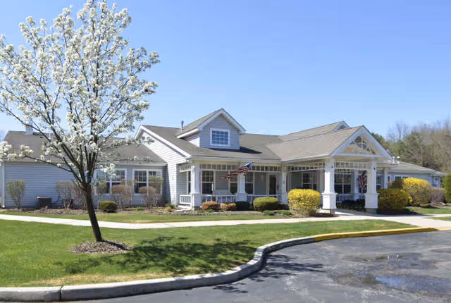 Single-story white senior living building with a covered entrance, American flags, and a flowering tree on a sunny day.