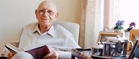 An elderly man wearing glasses and a light-colored shirt is sitting in a comfortable chair, holding an open book and smiling. Behind him, there is a window with light curtains and a table with a tea set and some small potted plants.