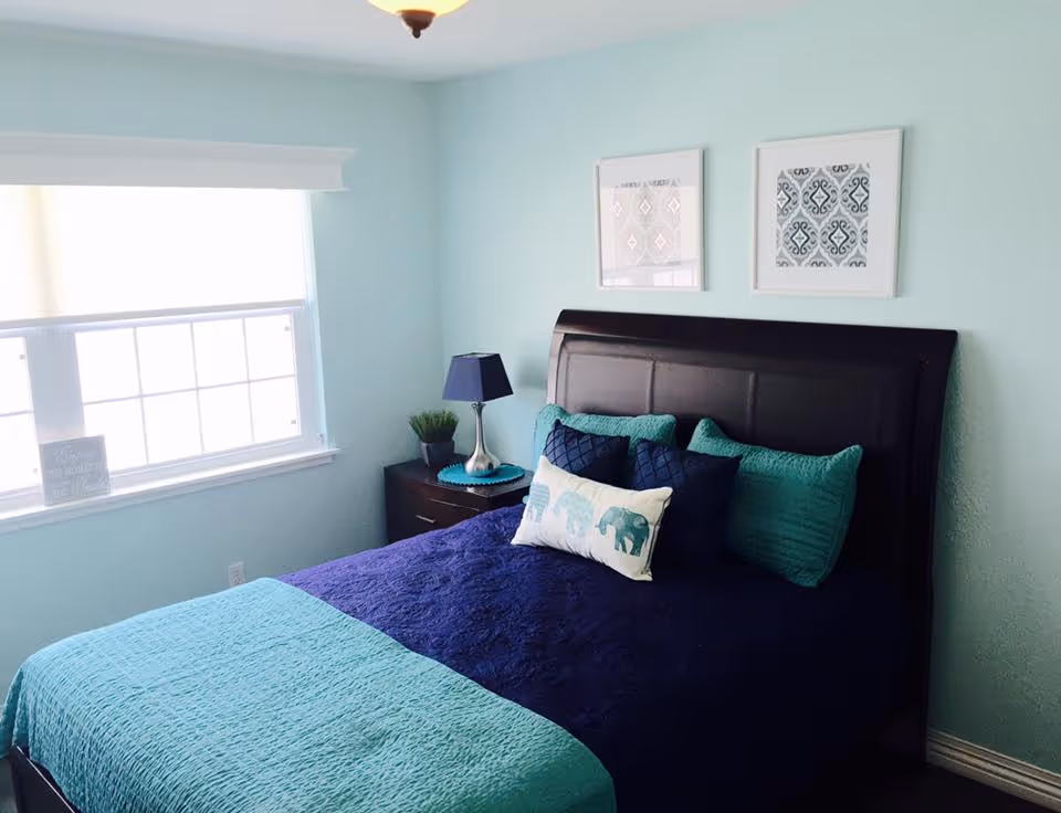 A bedroom with light blue walls and a large window with a white blind. The bed has a dark wooden headboard and is made with a navy blue and teal bedspread, decorated with teal and navy pillows, including one with an elephant design. There is a dark wooden nightstand next to the bed with a blue lamp and a small plant. Two framed patterned artworks hang above the bed.