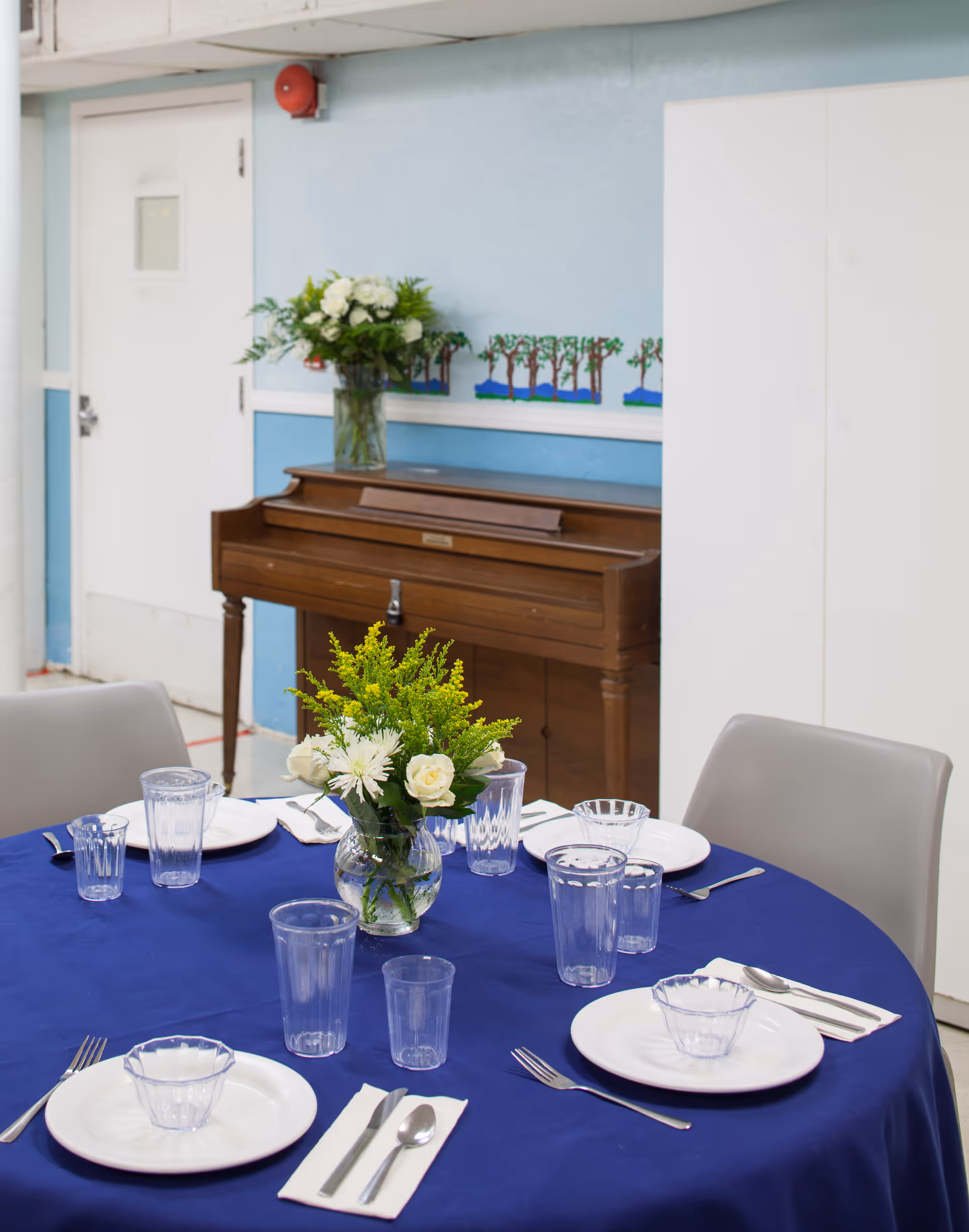 Round dining table with blue tablecloth, flower centerpiece, glassware, and a piano in the background.