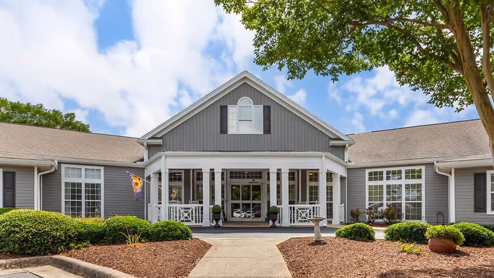 Front exterior view of a single-story building with gray siding and white trim, featuring a covered porch with white railings and two potted plants. The building is surrounded by neatly trimmed bushes and mulch beds, with a clear blue sky and some clouds in the background.