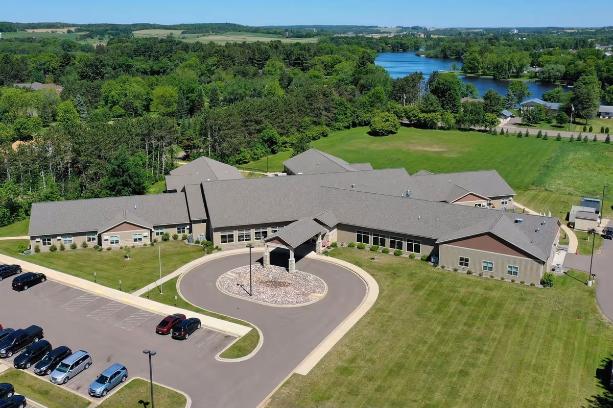Aerial view of a single-story senior living facility with a circular drive, parking lot and lawns, set among trees with a lake in the background.