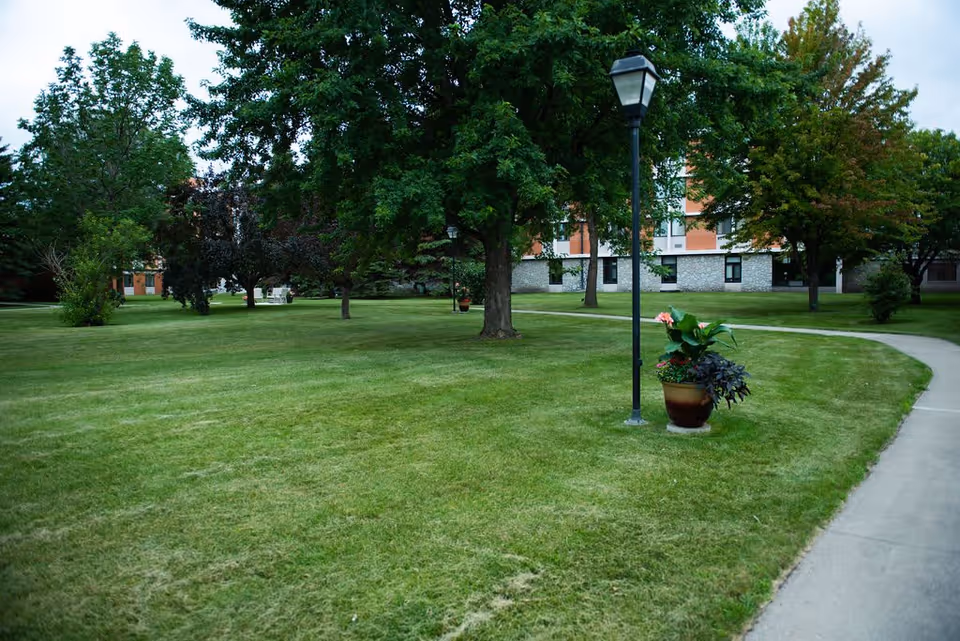A well-maintained outdoor garden area with green grass, several trees, a curved concrete pathway, a lamppost, and a flower pot with blooming plants. A multi-story building is visible in the background.