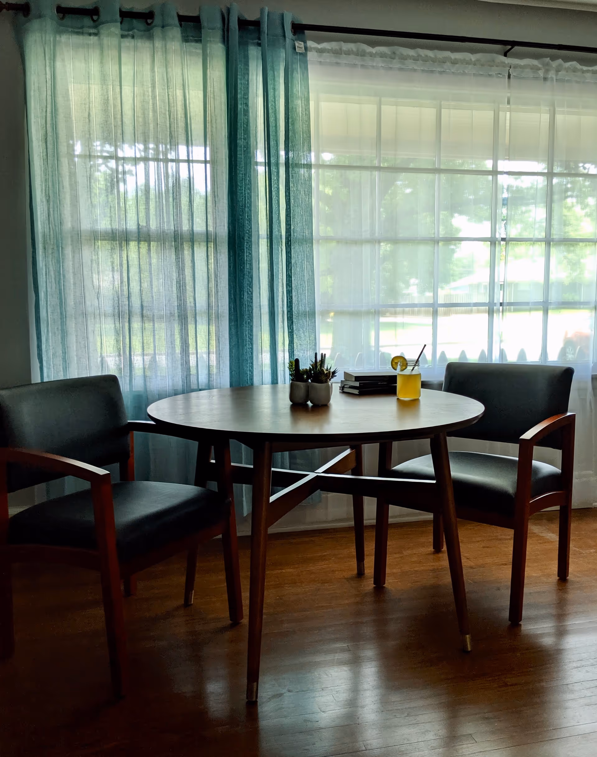 Round wooden table with two armchairs by a large window with sheer teal curtains, holding a small plant, books, and a yellow drink.