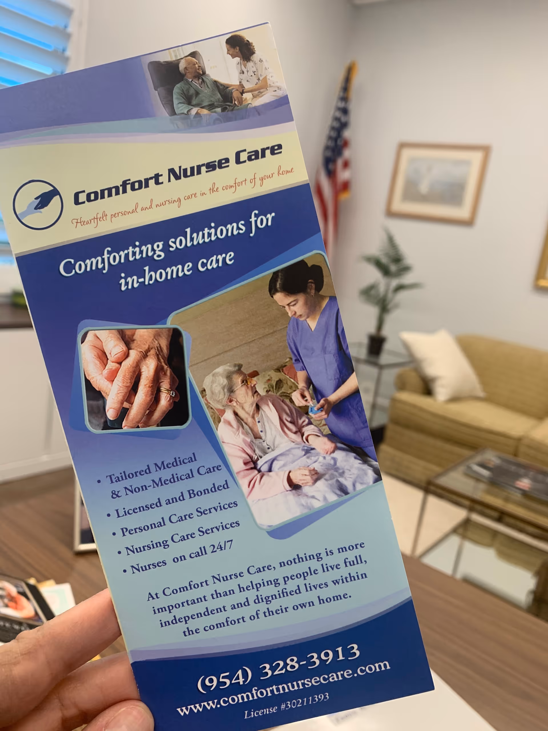 A Comfort Nurse Care brochure held in a living room with sofa and plant.