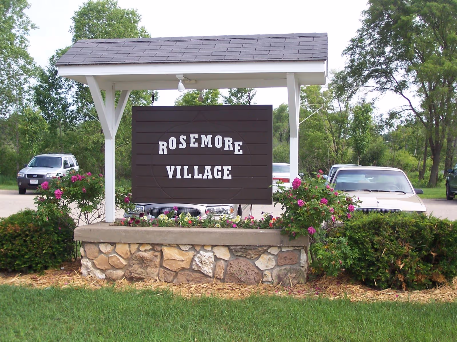 Exterior sign reading Rosemore Village, surrounded by flowers with parked cars and trees nearby.
