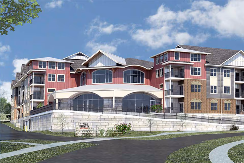Exterior view of a large multi-story assisted living facility building with red and beige siding, multiple windows, balconies, and a sloped roof under a partly cloudy blue sky. There is a stone retaining wall and a sign that reads Drumlin Reserve in front of the building.
