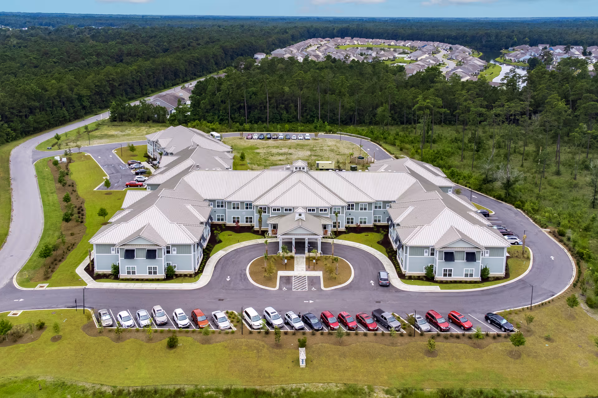 Aerial view of Arbor Landing at Surfside, a large senior living facility surrounded by greenery and trees, with multiple parking areas and a circular driveway at the entrance.