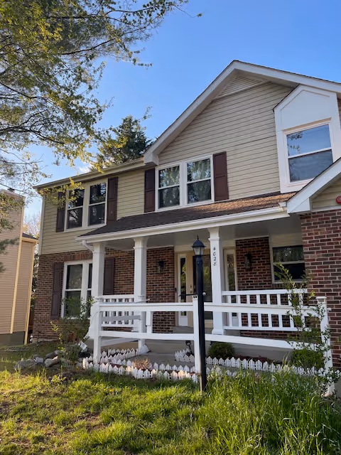 Two-story assisted living facility with beige siding, brick lower level, white porch, and front yard.