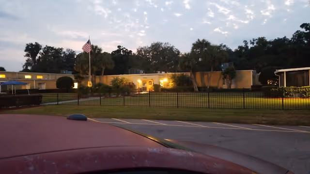 Exterior view of a single-story building at dusk with lights on inside. The building is surrounded by a lawn, palm trees, and a black metal fence. An American flag is visible on a flagpole near the center of the image. The sky is partly cloudy with the last light of the day.