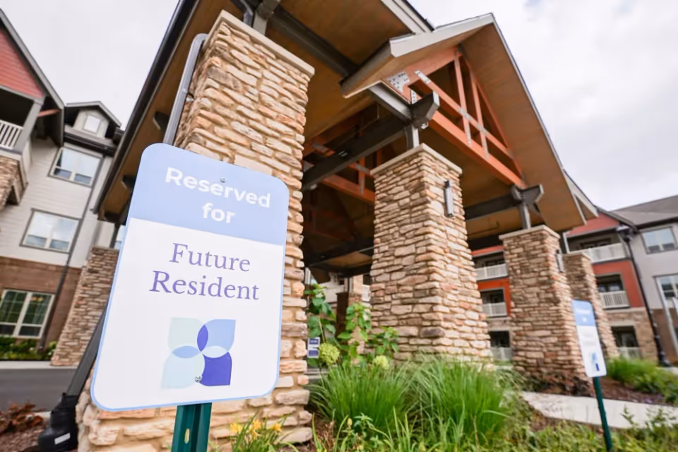 Entrance of a senior living facility with stone pillars and a wooden roof structure. A sign in the foreground reads 'Reserved for Future Resident'. The building has multiple windows and balconies.
