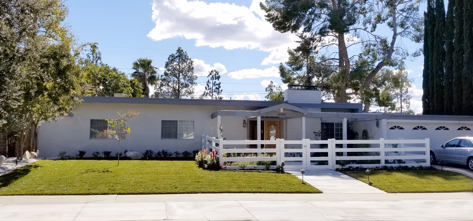 Single-story white senior living facility with a white fence, front entrance, and manicured lawn.