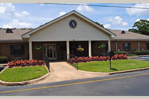 Front exterior view of a single-story brick building with a white gabled entrance supported by columns. There are flower beds with pink flowers and green grass on either side of the walkway leading to the entrance. A circular emblem with the letter 'A' is displayed at the peak of the gable. The sky is partly cloudy.
