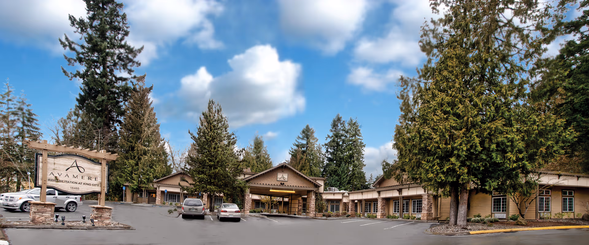 Front exterior of the Avamere Rehabilitation of King City building with a parking lot, entrance canopy, sign, and surrounding trees under a blue sky.