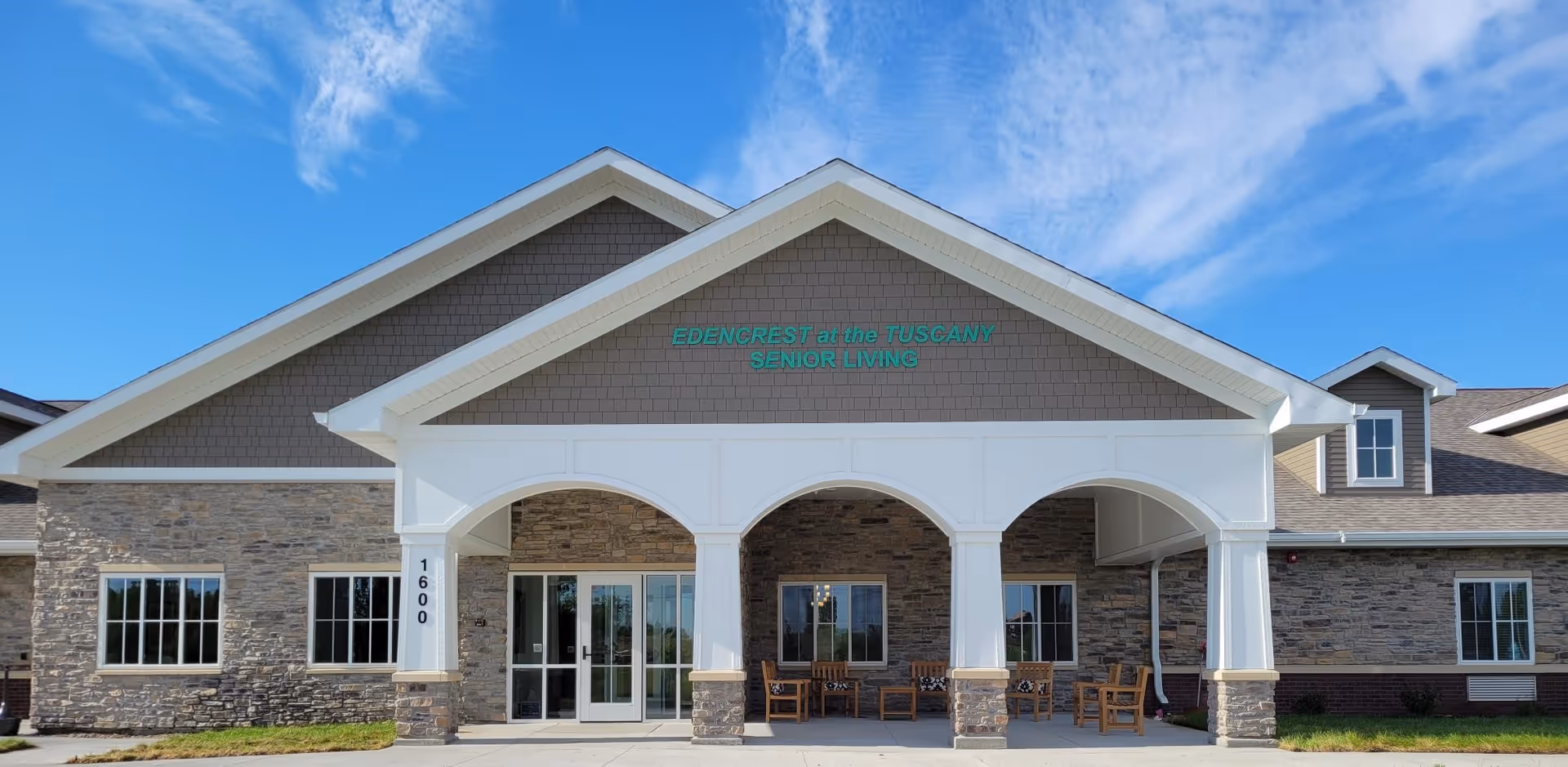 Front entrance of Edencrest at the Tuscany senior living building featuring a covered portico with stone facade and outdoor seating.