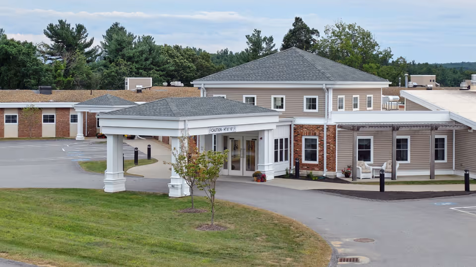 Exterior view of Holden Rehabilitation & Skilled Nursing Center showing the main entrance with a covered drop-off area, surrounded by a driveway and green lawn, with trees in the background under a cloudy sky.