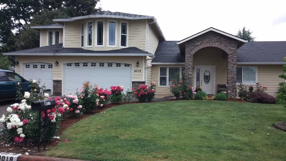 Front exterior view of a two-story residential building with beige siding, a stone archway entrance, a white front door with an oval window, and a three-car garage. The well-maintained lawn is bordered by flower beds filled with blooming pink and white flowers. A black mailbox is visible near the driveway.