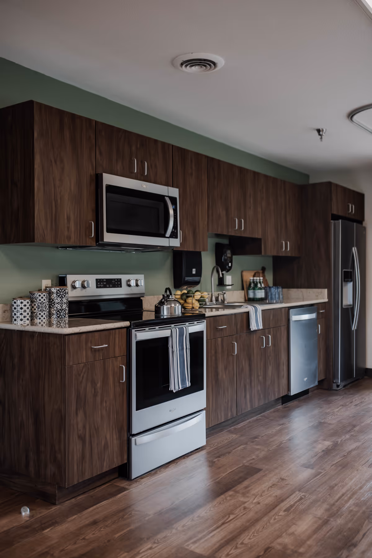 Modern kitchen with dark wood cabinets, stainless steel appliances including a microwave, stove, dishwasher, and refrigerator. The countertop has decorative containers, a kettle on the stove, a bowl of bananas, and some glasses near the sink. The floor is wooden and the walls are painted green.