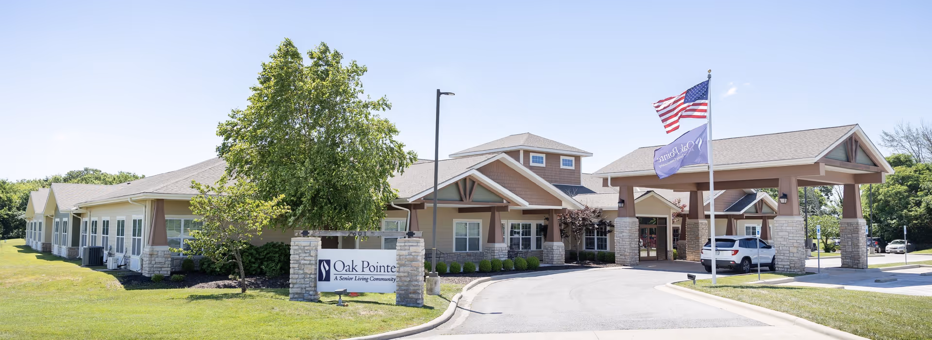 Exterior view of Oak Pointe of Neosho senior living community building with a covered entrance, an American flag and a community flag flying on flagpoles, surrounded by green grass and trees under a clear blue sky.