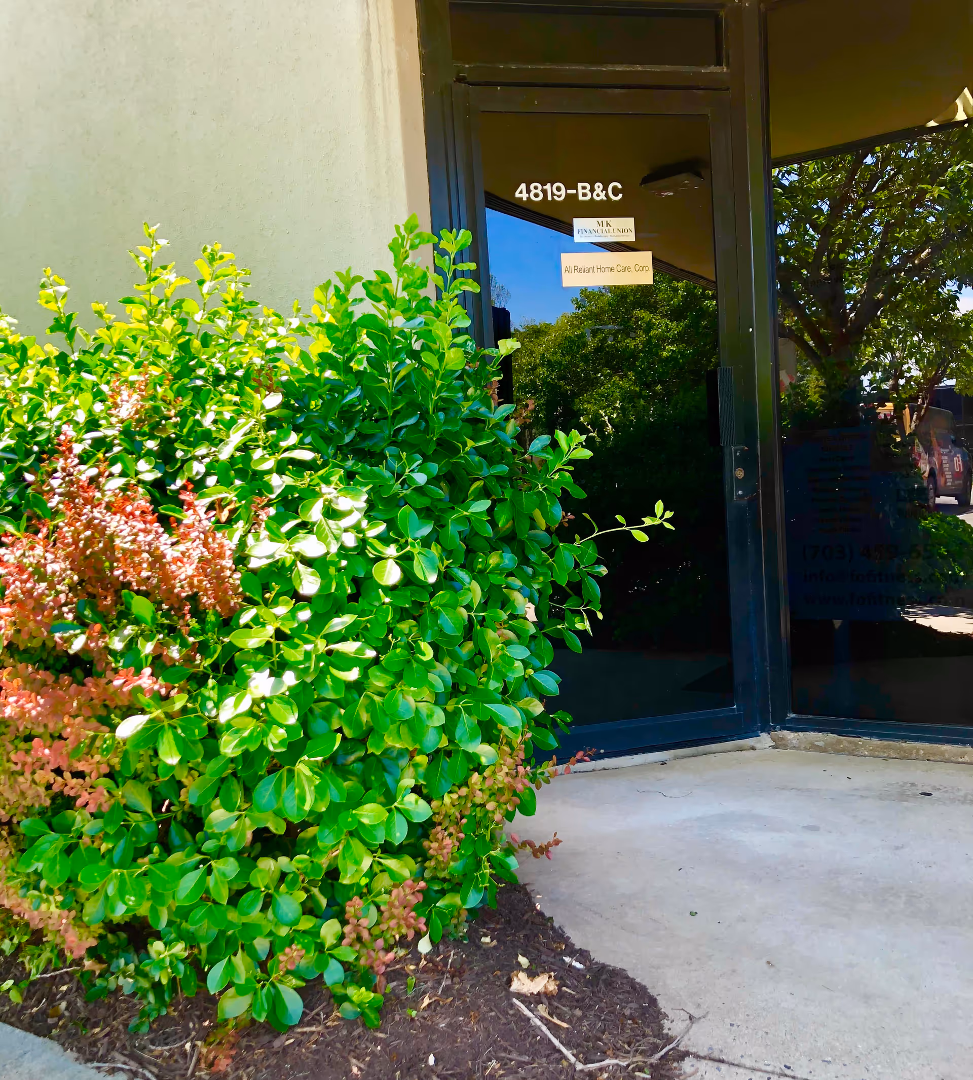 Glass entrance door of All Reliant Home Care Corp with lush shrubs outside.