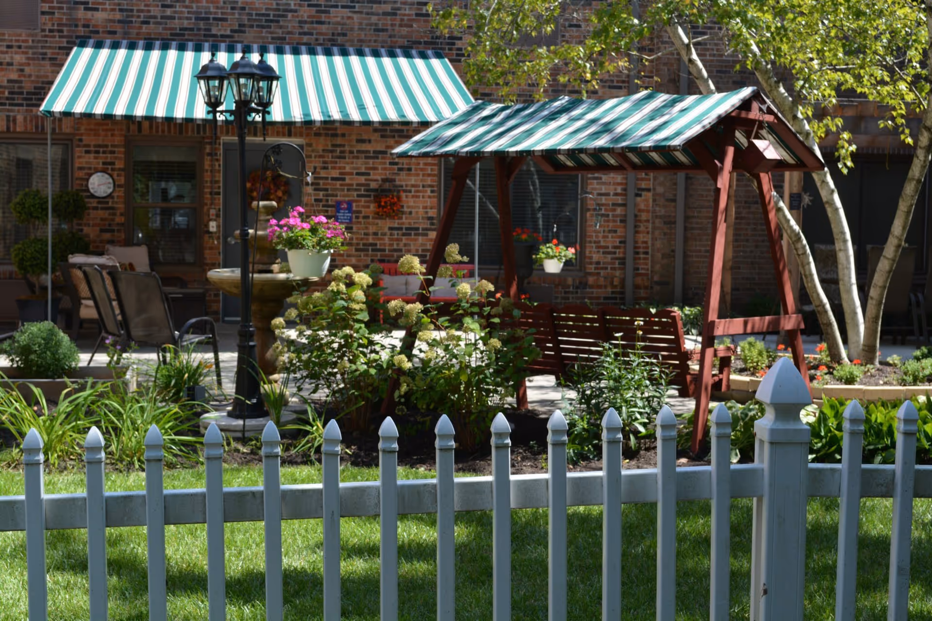 Outdoor garden area with green grass, a white picket fence in the foreground, a wooden swing with a green and white striped canopy, potted flowers, shrubs, and a brick building with a matching green and white striped awning in the background.