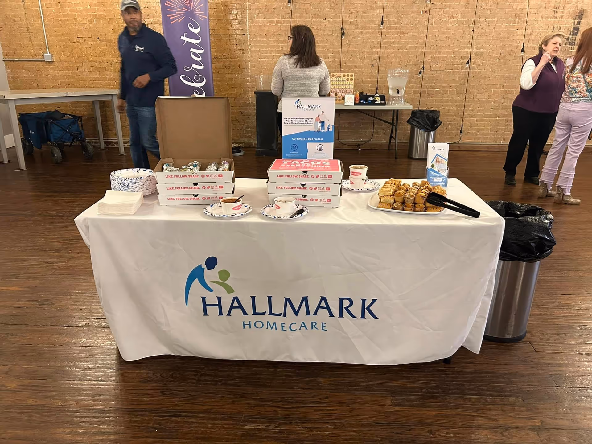 Indoor event table with Hallmark Homecare banner and snacks, brick wall background, several people.