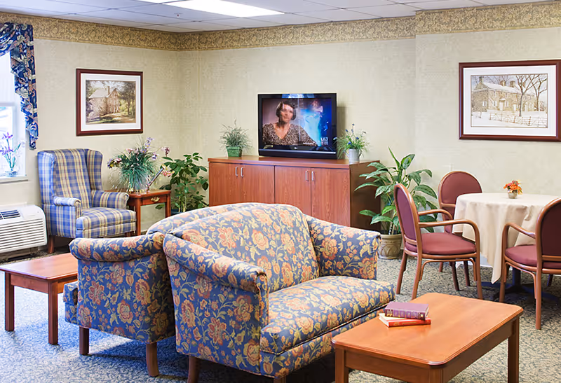 A cozy living room area in a senior living facility with floral patterned sofas and armchairs, a wooden coffee table with books, a TV on a wooden cabinet, potted plants, a round table with four chairs, framed artwork on the walls, and a window with floral curtains.