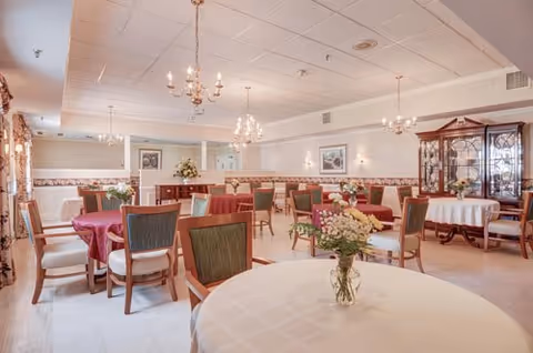 A spacious senior living dining room with round tables, chandeliers, and floral centerpieces.