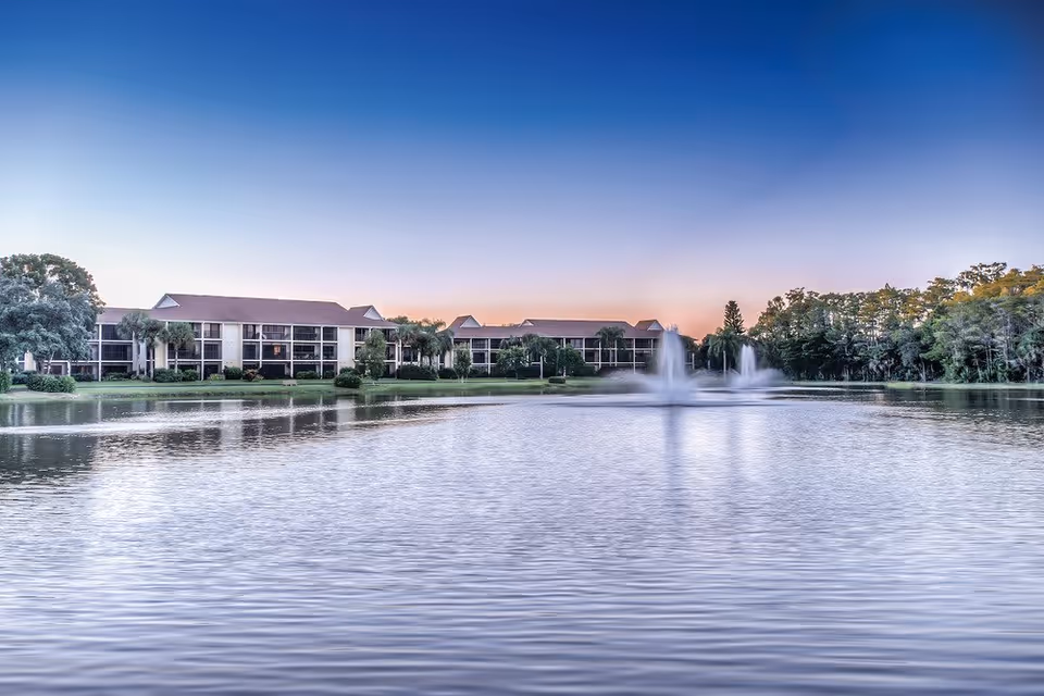 View of a senior living facility named Vi at Bentley Village across a large pond with two water fountains, surrounded by trees and greenery under a clear sky at sunset.