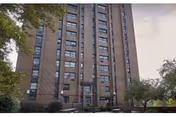 Tall brick building facade with many windows, trees, and a sidewalk.