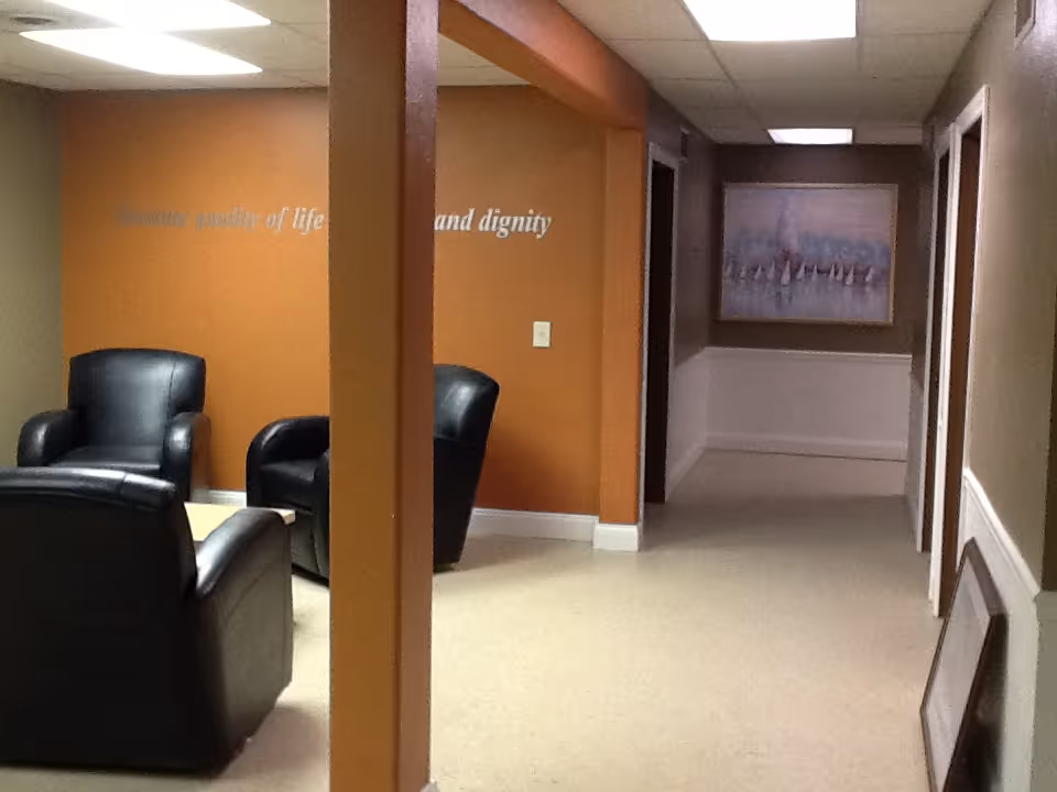 Interior hallway with an orange wall, black chairs, and framed artwork along a waiting area.