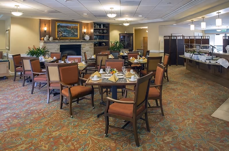 A dining room with multiple set tables and upholstered chairs, a buffet station to the right, and a fireplace with a framed painting at the back.