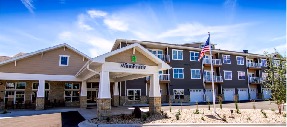 Exterior view of the Winn Prairie senior living facility showing a multi-story building with balconies, an entrance canopy supported by stone pillars, an American flag on a flagpole, and a clear blue sky.