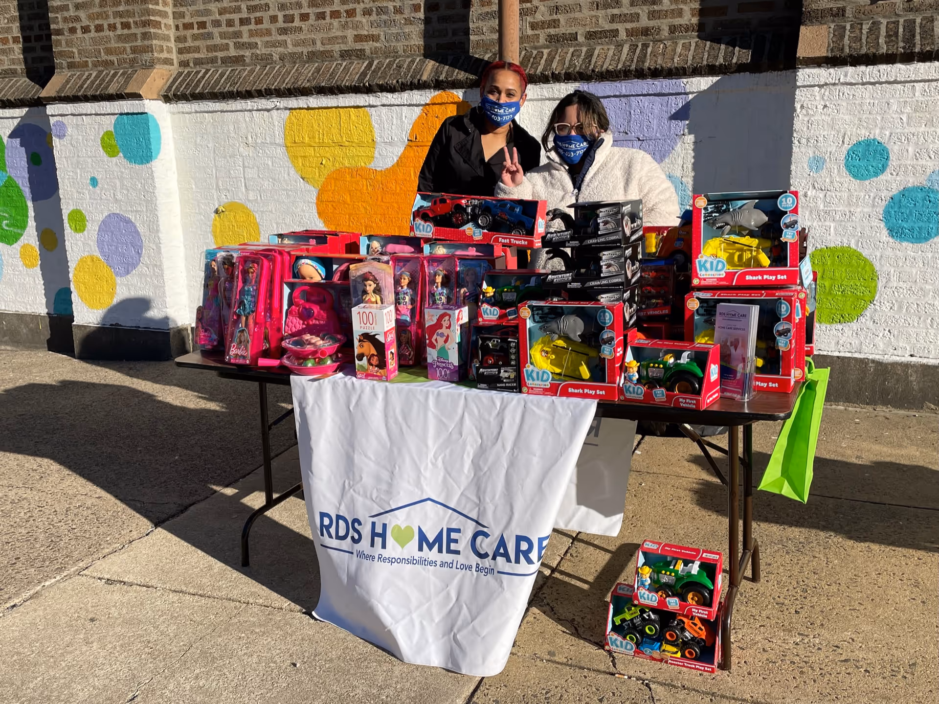 Two masked people stand behind a table of toys outside a colorful mural.